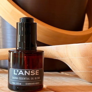 May include: A small amber glass bottle with a black dropper cap containing L'ANSE Sauna Essential Oil Blend. The label displays the text 15mL | 0.5 oz and STARBOARD NORTH. The bottle is positioned in front of a wooden sauna ladle and bucket.