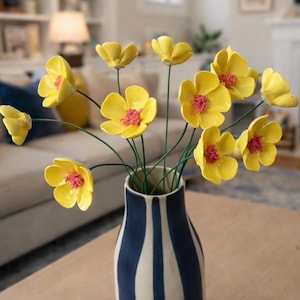 Puede incluir: Un jarrón de flores amarillas con centros rosas y tallos verdes. Las flores están en un jarrón a rayas azules y blancas. El jarrón está sobre una mesa de madera en un salón.