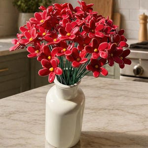 以下が含まれることがあります： 白いセラミックの花瓶に入った、黄色の中心を持つ鮮やかな赤い花のブーケ。 花は光沢のある仕上がりで、緑色の茎で配置されています。 花瓶は円筒形で、上部がわずかにフレアしており、大理石のカウンタートップに置かれています。