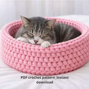 May include: A grey tabby cat sleeps peacefully in a round, pink, crocheted cat bed. The bed is made of thick yarn and has a textured appearance. The text "PDF crochet pattern_instant download" is at the bottom of the image.