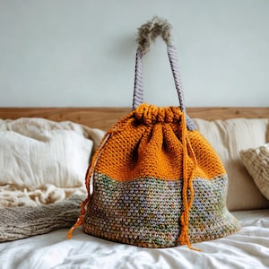 May include: A handmade, orange and gray drawstring bag with a textured knit design. The bag features a gray handle and a fluffy, gray pom-pom detail at the top. The bag is sitting on a white bedspread.
