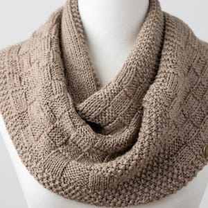 May include: A tan-colored, knitted cowl scarf with a textured, geometric pattern. The scarf is displayed on a white mannequin and features a button closure. The knitted design is visible, showcasing the craftsmanship of the piece.