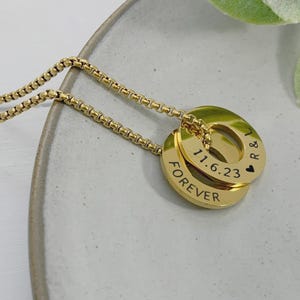 May include: A gold-colored necklace with two circular pendants. The top pendant has the date "11.6.23" and a heart symbol. The bottom pendant has the word "FOREVER" engraved. The necklace is on a light gray plate with a sprig of green leaves.