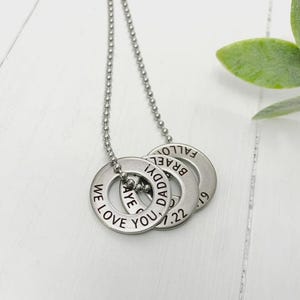 May include: A silver-colored necklace with three circular pendants. Each pendant has engraved text: "WE LOVE YOU," "DADDI!," and a name and date. The necklace hangs on a delicate chain, perfect for a personalized gift.