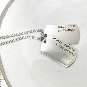 May include: Two brushed silver dog tag necklaces with engraved names and dates. The tags are connected by a silver chain and rest on a white ceramic plate. The tags read "Jaxon Cole IV.XV.MMXX" and "Hunter Jameson X.XXV.MMXXI".
