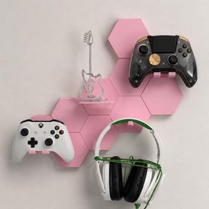 May include: A set of pink hexagonal wall-mounted shelves holding a black and gold game controller, a white game controller, a silver wire guitar sculpture, and a pair of white and green over-ear headphones hanging from a small hook.