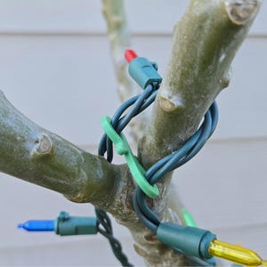 May include: Close-up of a tree branch with green and blue Christmas lights wrapped around it. The lights have different colored bulbs, including red, blue, and yellow. A green clip secures the lights to the branch.