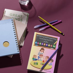 Pode incluir: Uma coleção de material escolar sobre uma superfície roxa. Inclui cadernos, canetas, lápis e um livro intitulado "Identifying and Supporting Learning Barriers in the Classroom". Um copo de água também está presente.