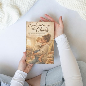 May include: A book titled "Embracing the Chaos" is held by a person. The cover features a watercolor illustration of a mother embracing a child. The book's subtitle reads "Celebrating Motherhood & Finding Joy in the Journey."
