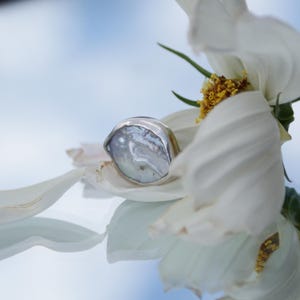 Peut inclure: Une bague ornée d'une perle irrégulière et irisée de couleur claire, sertie dans un anneau en métal argenté lisse et poli. La bague est nichée parmi les pétales blancs d'une fleur au cœur jaune, se reflétant sur une surface lisse.