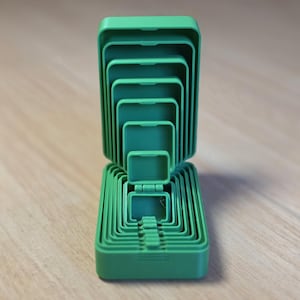 May include: A set of nested, green plastic containers with hinged lids, arranged in a stacked configuration. The containers vary in size, with the smallest at the center and the largest at the base. The lids are open, revealing the interior of each container.