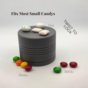 May include: A gray cylindrical container with the text "Fits Most Small Candies" and "Twist to Lock." The container holds gum and is surrounded by Skittles and M&Ms candies. The container is designed to hold small candies.