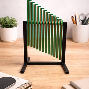 May include: A decorative desktop musical instrument with green and orange vertical bars. The bars are arranged in a descending order and are supported by a black metal frame. The instrument sits on a wooden desk, with a notebook and a plant in the background.