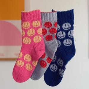 Puede incluir: Tres pares de calcetines de punto en rosa, gris y azul, cada uno con un estampado de caras sonrientes repetidas. Los calcetines rosas tienen caras amarillas, los calcetines grises tienen caras rojas y los calcetines azules tienen caras blancas. Los calcetines cuelgan frente a un fondo neutro.