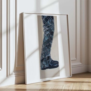 Peut inclure: Une œuvre d'art encadrée représentant une jambe vêtue d'un jean en denim déchiré et de bottes noires. L'œuvre est placée sur un fond neutre et encadrée d'une bordure blanche, appuyée contre un mur.