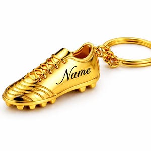 Op de afbeelding: Een goudkleurige voetbalschoen sleutelhanger met het woord "Name" in zwart schrift. De sleutelhanger heeft een ring en ketting bevestiging. De schoen heeft veters en noppen.