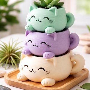 Könnte beinhalten: Drei gestapelte, katzenförmige Blumentöpfe in Pastellfarben: Mintgrün, Lavendel und Creme. Jeder Topf hat ein lächelndes Katzengesicht und eine kleine Sukkulentenpflanze. Die Töpfe stehen auf einem Holztablett und bilden eine verspielte Dekoration.