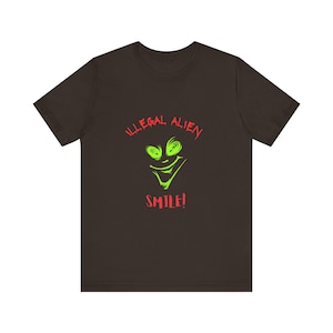 May include: A dark brown short-sleeve t-shirt featuring a graphic of a bright green alien face with a wide grin. The text "ILLEGAL ALIEN" is printed in red above the face, and the word "SMILE!" is printed in red below the alien graphic.