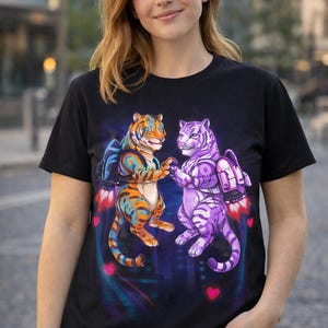 以下が含まれることがあります： 黒いTシャツ。ジェットパックを装着し、手をつないでいる2匹のトラのカラフルなグラフィック。1匹はオレンジと青、もう1匹は紫です。デザインにはハートと都市景観の背景が含まれています。