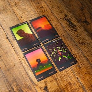 May include: Four Instax film cards with colorful sunset and silhouette imagery. The cards have black borders and the text "FUJIFILM INSTAX" and "INSTAX" printed on them. The cards are arranged on a wooden surface.