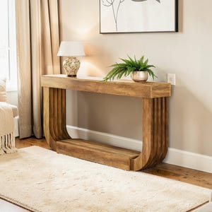May include: A light brown wooden console table with a curved design. The table has a rectangular top and a lower shelf. A white lamp with a floral design and a small potted plant sit on top of the table. The table is 60 inches long.