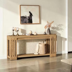 May include: A light brown wooden console table with a lower shelf and vertical slat detailing on the sides. The table is decorated with a painting, vases, candles, and a woven basket.