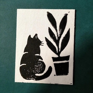 May include: Black and white linocut print featuring a cat and a potted plant. The cat is seated, facing the plant with its tail curled. The plant has large, stylized leaves. The print is on a white rectangular paper.
