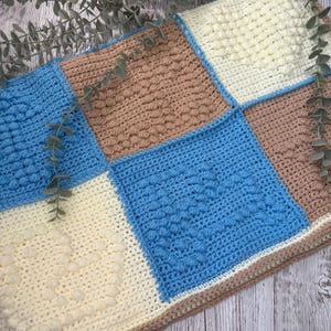 May include: A handmade crochet blanket featuring a patchwork design. The blanket showcases squares of light blue, cream, and tan, with textured stitch patterns. The blanket is a cozy, handcrafted item.