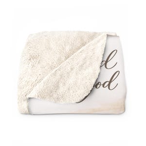 May include: A cream-colored sherpa blanket with the words "Good God" in a brown script font. The blanket is folded, revealing the soft, fluffy texture of the sherpa material. The background is white.