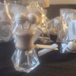 Pu&ograve; includere: Profumatore per auto in vetro trasparente con parte superiore in legno e due accenti a sfera in legno. Diversi profumatori simili sono visibili in sacchetti di plastica. L'etichetta sulla busta recita "CRANBERRY".