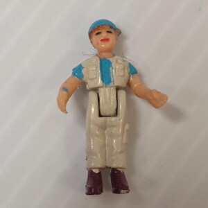 Pode incluir: Pequena figura de plástico de uma pessoa usando um boné azul, um colete branco com detalhes azuis e calças brancas. A figura tem botas marrons e está de pé com os braços estendidos.