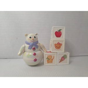May include: A pearlescent snowman figurine with a purple scarf and pink buttons, next to a stack of white toy blocks. The blocks feature illustrations of an apple, a teddy bear, a cat, and an ice cream cone.