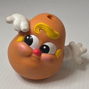 May include: A small, orange, potato-shaped plastic figurine with a smooth surface. It features large white eyes with black pupils, a small red nose, pink cheeks, and yellow hair. Two white, four-fingered hands extend from the sides of the body.