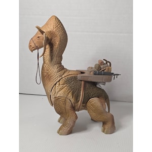 Op de afbeelding: Tan-kleurige Bantha speelgoedfiguur met een zadel en accessoires. De figuur heeft een lange nek, grote kop en gedetailleerde getextureerde huid. Het zadel bevat diverse items, waaronder een blastergeweer en opgerolde items.