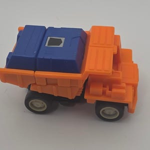 May include: A small orange plastic toy dump truck with a blue cargo bed. The truck features black wheels with light gray centers and a small silver and black emblem on the top of the blue cargo section. The design is blocky and angular.