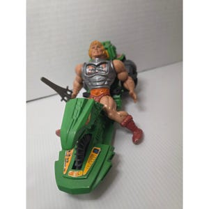May include: A vintage action figure of He-Man riding a green vehicle. The figure has blonde hair, a silver chest plate, and red shorts. The vehicle is green with yellow accents and a detailed design. A black sword is held by the figure.