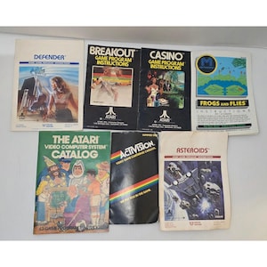 Puede incluir: Una colección de manuales y catálogos de videojuegos Atari antiguos. Los títulos incluyen "Defender", "Breakout", "Casino", "Frogs and Flies", "The Atari Video Computer System Catalog", "Activision" y "Asteroids". Los manuales presentan gráficos y texto coloridos.