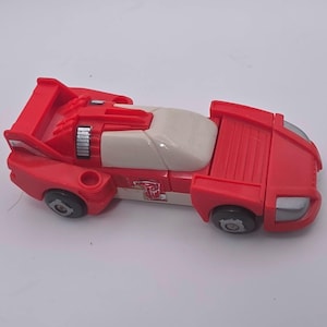 May include: A red and white toy race car with a spoiler, black wheels, and a silver front bumper. The vehicle features a white cockpit area and a small red and white emblem on the side panel, resembling a stylized robot face icon on the door.