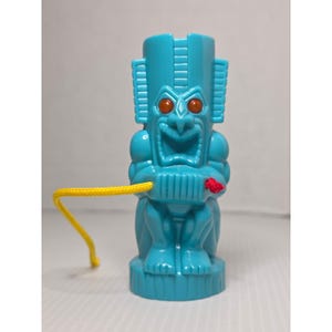 Puede incluir: Una figura de plástico de color turquesa brillante estilo tiki con un rostro expresivo de boca ancha y ojos naranjas. La figura tiene un cordón amarillo que sale de un lado y un pequeño detalle de nudo rojo al frente, sobre una base cilíndrica.