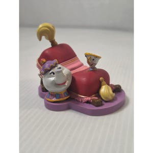 May include: A decorative figurine featuring characters from a fairy tale. The figurine includes a red hat with a yellow plume, a white teapot with a purple hat, and a small teacup. The base is purple.