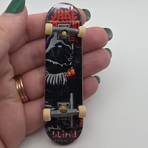 Peut inclure: Un skateboard miniature pour les doigts avec un motif représentant une silhouette encapuchonnée aux yeux brillants. Le plateau porte les inscriptions Jake Brown et Blind. Il possède des trucks en métal gris et quatre petites roues blanc cassé.