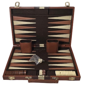 May include: Open backgammon set in a brown leather case. The interior features a playing surface with alternating tan, brown, and white triangles. Includes brown leather dice cups, dice, and game pieces. The case is closed with a metal clasp.