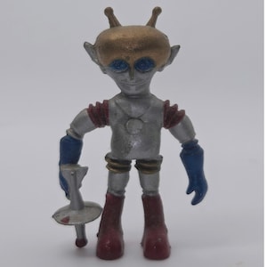May include: A small alien figurine with a metallic gold head, large blue eyes, and antennae. The body is silver with red shoulder accents, blue gloves, and dark red boots. The figure holds a silver, disc-shaped weapon in its right hand.