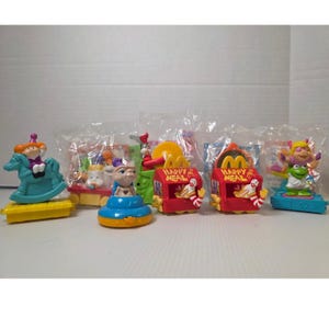 May include: A collection of vintage McDonald's Happy Meal toys. The toys feature characters on rocking horses, E.T., and Muppets. The toys are in their original packaging, with bright colors and detailed designs.