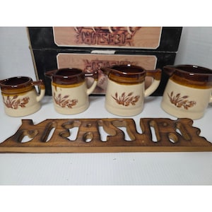 Puede incluir: Un juego de crema de cerámica vintage con cuatro cremeras y un salvamanteles de madera. Las cremeras son beige con detalles marrones y diseños florales. El salvamanteles de madera deletrea la palabra "JESUS".