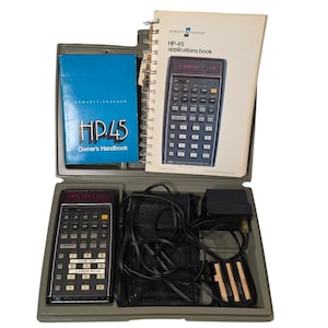 May include: A vintage Hewlett-Packard HP-45 calculator kit in a gray case. The kit includes the calculator, a blue owner's handbook, an applications book, a black power adapter, and a black carrying case. The calculator has numerous buttons and a red display.