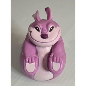 Könnte beinhalten: Eine lila Cartoon-Figur mit einem lächelnden Gesicht, großen Ohren und kleinen Armen und Füßen. Die Figur ist hauptsächlich lila mit helleren rosa Akzenten auf Gesicht, Ohren und Füßen. Die Figur hat schwarze Augen und eine dunkelviolette Nase.