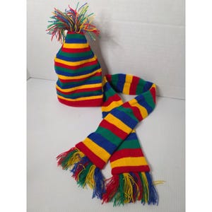 Può includere: Un set composto da un berretto in maglia e una sciarpa abbinata con righe orizzontali rosse, gialle, blu e verdi. Il berretto è sormontato da un pon-pon in filato multicolore e le estremità della sciarpa presentano lunghe frange multicolori.