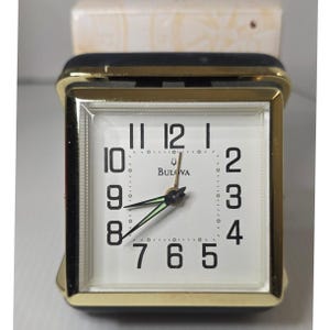 May include: A vintage Bulova alarm clock with a gold-tone frame and a white face. The clock features black numerals, black and green hands, and the Bulova logo. The clock is rectangular and has a black top.