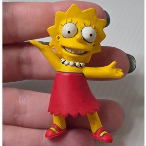 Op de afbeelding: Een klein geel Lisa Simpson-figuurtje met stekelig haar, grote witte ogen en een brede glimlach. Het personage draagt een rode jurk met een gekartelde zoom, een witte parelketting en rode schoenen met gele accenten, met de armen gespreid.